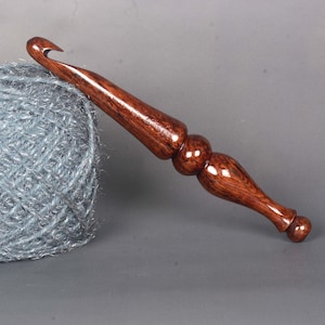 May include: A wooden crochet hook with a dark brown stain. The hook has a rounded handle and a pointed tip. The hook is lying on a ball of gray yarn.