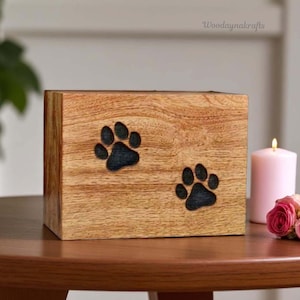 Puede incluir: Urna de madera para mascotas con dos huellas de patas negras grabadas. La urna es rectangular y de madera clara. El fondo incluye una vela encendida y un pequeño ramo de rosas rosas.