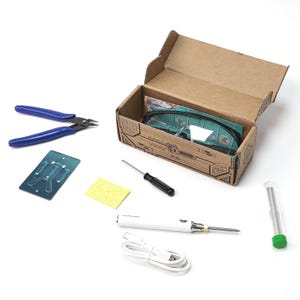 May include: A soldering kit featuring blue wire cutters, a soldering iron, safety glasses, a screwdriver, and a small sponge. The tools are arranged on a white surface, with some items inside a cardboard box.