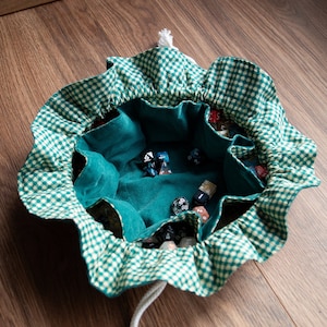 May include: A green and white gingham dice bag with a drawstring closure. The interior is a solid teal color and holds several multi-sided dice. The bag is open, displaying the dice inside.