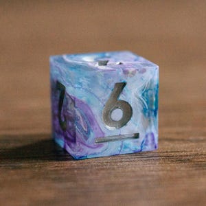 Puede incluir: Un dado de seis caras con un diseño veteado azul, morado y blanco. El número 6 se muestra en plata, con el número 7 visible en un lado adyacente. El dado está sobre un fondo de madera borroso.