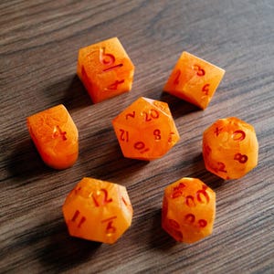 May include: Set of seven orange resin dice with red numbering, including a D20, D12, D10, D8, D6, D4, and a fractional die. The dice have a marbled appearance and are designed for tabletop role-playing games.