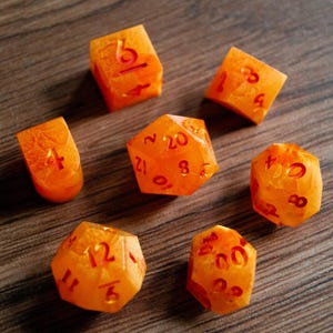 May include: A collection of orange dice with red numbering, including a D20, D6, and D4. The dice are arranged on a wooden surface. The dice have a textured appearance.