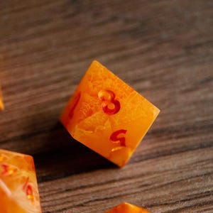 May include: Close-up of a six-sided die with the number 3 prominently displayed. The die is a translucent orange color with red numbering and a textured interior, resting on a wooden surface.