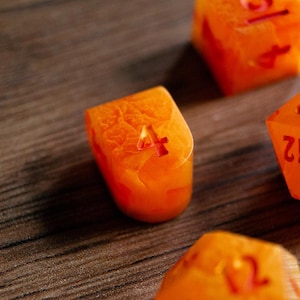 May include: Close-up of orange resin dice with red numbering. The dice are translucent, with a textured, marbled appearance. The number "4" is visible on the face of one die. Other dice with visible numbers are also in the image.