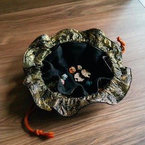 Puede incluir: Bolsa de dados negra y dorada con un diseño de telaraña. La bolsa tiene un interior negro y un cordón naranja. Varios dados de varios colores están dentro de la bolsa. La bolsa está sobre una superficie de madera.