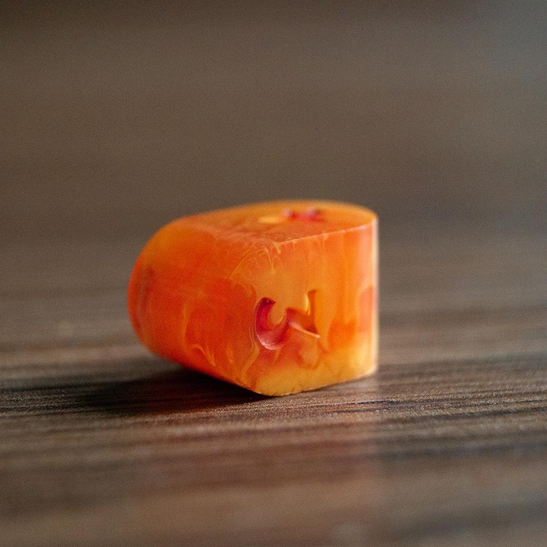 May include: Close-up of an orange and yellow resin object with a carved "W" on its side. The object has a rounded top and a flat bottom, with a marbled appearance. The background is a blurred wooden surface.