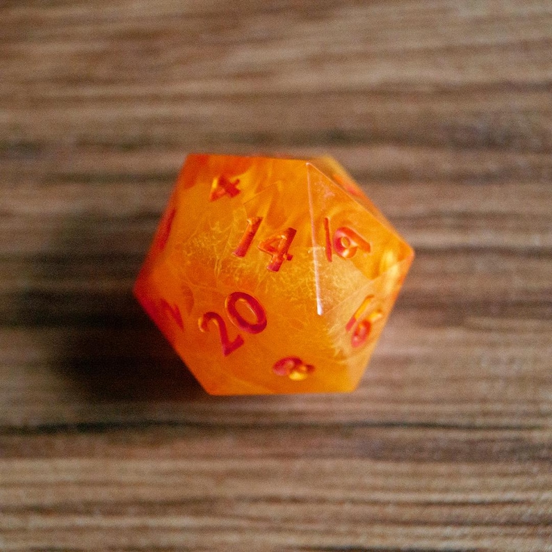 May include: A twenty-sided die with a fiery orange and red color scheme. The die is transparent, revealing an internal pattern. The numbers are in a bold, red font. This gaming accessory is perfect for tabletop games.