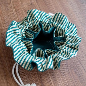 Peut inclure: Une pochette à cordon de serrage bleu sarcelle avec une doublure rayée blanche et verte. La pochette a un haut froncé et est ouverte pour révéler l'intérieur.