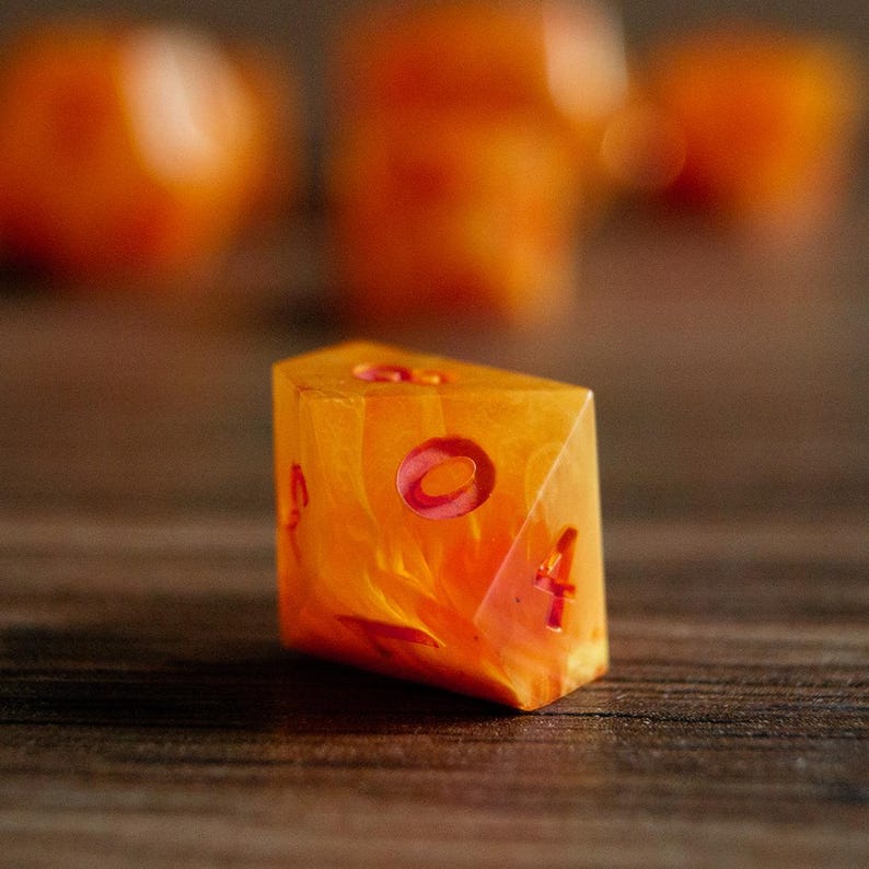 May include: A close-up of a single, orange and yellow marbled resin die with red numbering. The die is a unique shape, with one side angled. The background is blurred, with other dice visible.