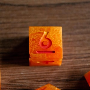 May include: Orange and red resin die with the number 6 etched in red. The die has a marbled appearance with a mix of orange and red hues. The die is a cube shape and is likely used for tabletop games.