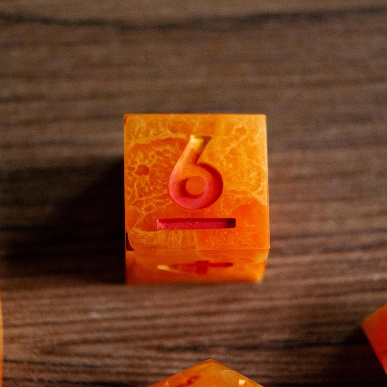May include: Orange and red resin die with the number 6 etched in red. The die has a marbled appearance with a mix of orange and red hues. The die is a cube shape and is likely used for tabletop games.