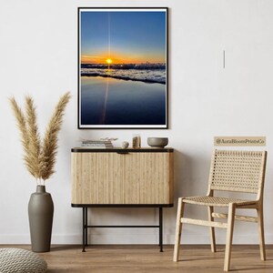May include: A framed print of a beach sunset. The sky transitions from blue to orange as the sun sets over the ocean. The water reflects the sky's colors. The print is displayed above a wooden cabinet with a vase of pampas grass.