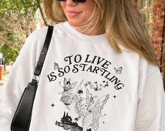 Peut inclure: Sweat-shirt blanc avec un motif graphique noir et du texte. Le motif présente des illustrations de fées et la citation "TO LIVE IS SO STARTLING. It leaves little time for anything else." d'Emily Dickinson.