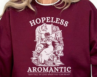 May include: Burgundy sweatshirt with a white graphic of a person reading in a pile of books. The text on the sweatshirt reads "Hopeless Aromantic Why fall in love when you can read?"