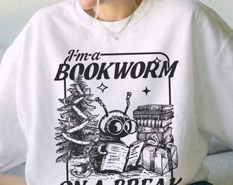 May include: White sweatshirt with a black and white graphic. The graphic features a cartoon bookworm with a Christmas tree, books, and gifts. Text on the sweatshirt reads "I'm a BOOKWORM ON A BREAK".