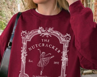 May include: A cardinal red crewneck sweatshirt featuring a white graphic print of a ballerina inside an ornate rectangular frame. The text reads THE NUTCRACKER, Est. 1892, and An enchanted Christmas performance. The design is centered on the chest.