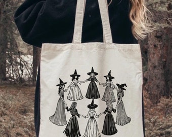 May include: Cream-colored tote bag featuring a black and white illustration of witches in a circle, holding hands. The text "GIRLS WILL BE GIRLS" is printed below the illustration. The bag has two handles and is held by a person.