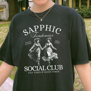 May include: Black t-shirt with a white graphic design that says "Sapphic Tendencies" with two women in dresses walking. The graphic also says "No Boys Allowed" and "Meets Weekly" and "Social Club Gay Vibes & Good Times".