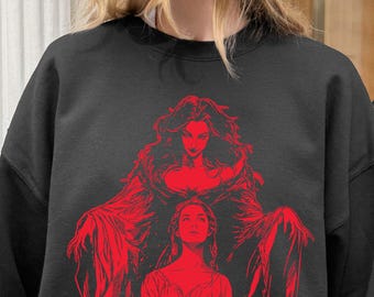 May include: A black crewneck sweatshirt featuring a bold red graphic print on the chest. The design depicts a central figure standing behind a seated person, both rendered in a detailed, illustrative style with flowing garments and long hair.