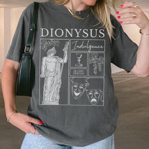 May include: Dark gray t-shirt with a graphic design featuring the word "DIONYSUS" above the word "Indulgence". The design includes illustrations of a statue, a wine glass, grapes, and theater masks. The phrase "IN VINO VERITAS" is also present.