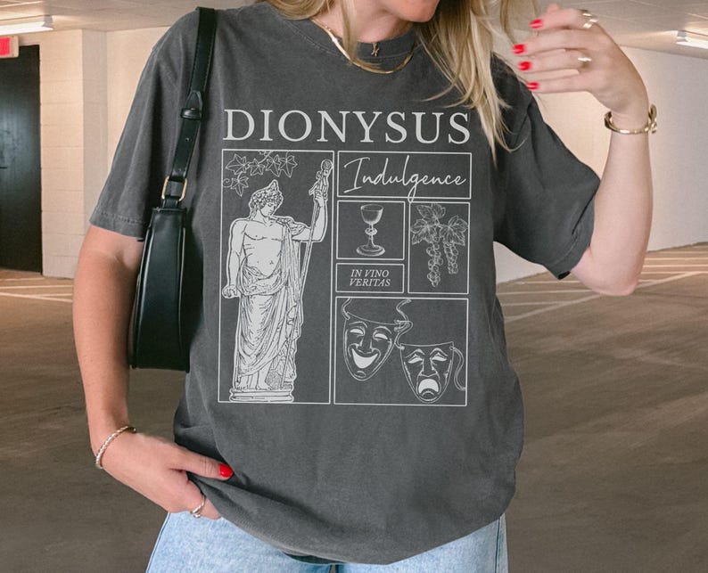 May include: Dark gray t-shirt with a graphic design featuring the word "DIONYSUS" above the word "Indulgence". The design includes illustrations of a statue, a wine glass, grapes, and theater masks. The phrase "IN VINO VERITAS" is also present.