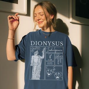 May include: A navy blue t-shirt with a white graphic design featuring the word "DIONYSUS" and the word "Indulgence". The design includes a statue, a wine glass, grapes, and theater masks.