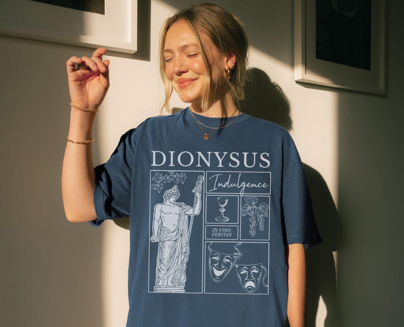 May include: A navy blue t-shirt with a white graphic design featuring the word "DIONYSUS" and the word "Indulgence". The design includes a statue, a wine glass, grapes, and theater masks.