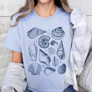 May include: A light blue t-shirt with a graphic print of various seashells in shades of blue. The shells are arranged in a random pattern on the shirt.