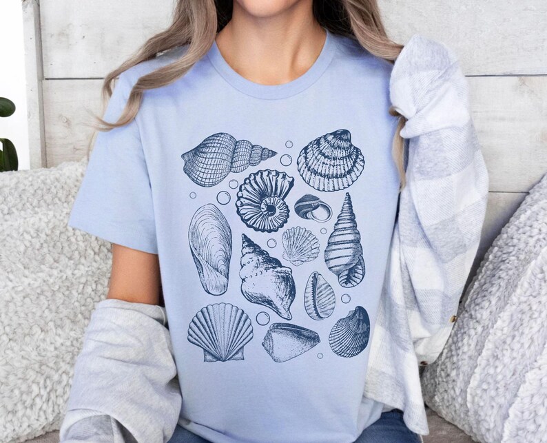 May include: A light blue t-shirt with a graphic print of various seashells in shades of blue. The shells are arranged in a random pattern on the shirt.