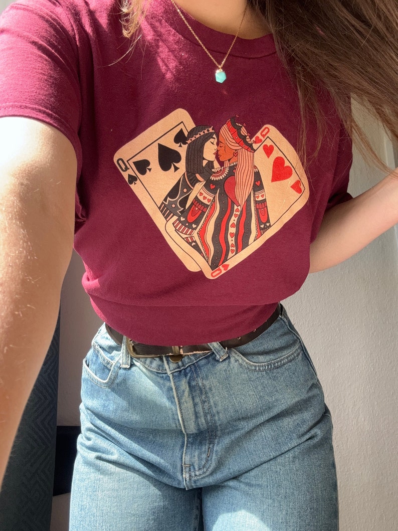 K&ouml;nnte beinhalten: Ein maroonfarbenes T-Shirt mit einem Aufdruck von zwei K&ouml;niginnen aus einem Kartenspiel, die sich k&uuml;ssen. Die K&ouml;niginnen tragen Kronen und halten sich an den H&auml;nden. Das T-Shirt wird von einer Person mit langen braunen Haaren und einer goldenen Halskette mit einem t&uuml;rkisfarbenen Anh&auml;nger getragen.