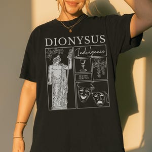 May include: Black t-shirt with a white graphic featuring the word "DIONYSUS" above the word "Indulgence". The graphic includes an illustration of a statue, a wine glass, grapes, and theater masks. The phrase "IN VINO VERITAS" is also present.