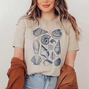 May include: A beige t-shirt with a blue and white print of various seashells. The print includes a variety of seashells, including a conch shell, a scallop shell, and a nautilus shell.