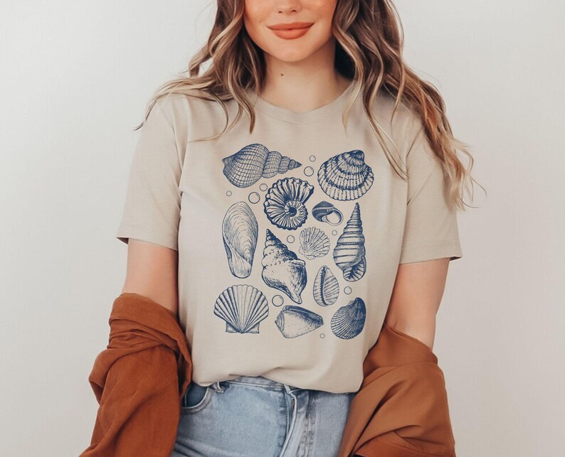 May include: A beige t-shirt with a blue and white print of various seashells. The print includes a variety of seashells, including a conch shell, a scallop shell, and a nautilus shell.