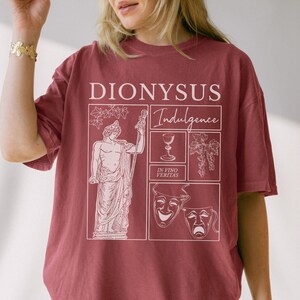May include: A maroon t-shirt featuring a white graphic design with the word "DIONYSUS" at the top. The design includes a statue, a wine glass, grapes, and theater masks, with the word "Indulgence" and the phrase "In Vino Veritas".