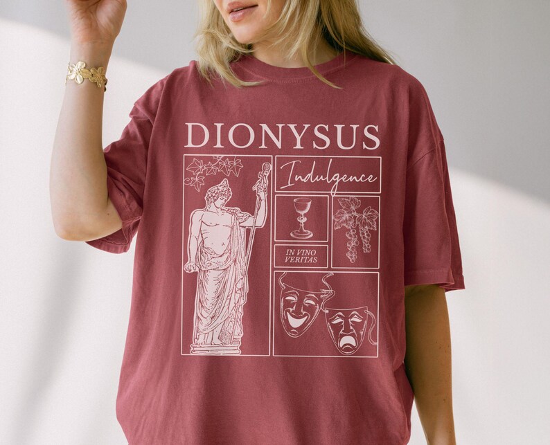 May include: A maroon t-shirt featuring a white graphic design with the word "DIONYSUS" at the top. The design includes a statue, a wine glass, grapes, and theater masks, with the word "Indulgence" and the phrase "In Vino Veritas".
