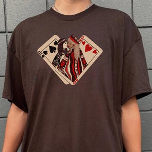 K&ouml;nnte beinhalten: Ein braunes T-Shirt mit einem Aufdruck von zwei K&ouml;niginnen aus einem Kartenspiel, die sich k&uuml;ssen. Die Herzk&ouml;nigin liegt auf der Pik-K&ouml;nigin. Der Aufdruck ist in Rot, Schwarz und Wei&szlig;.