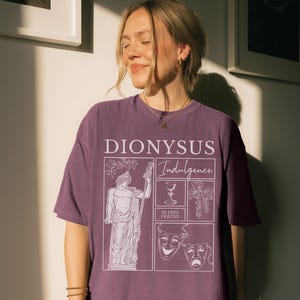 May include: A mauve-colored t-shirt with a graphic design featuring the word "DIONYSUS" and the word "Indulgence". The design includes a statue, a wine glass, grapes, and theatrical masks. The phrase "IN VINO VERITAS" is also included.