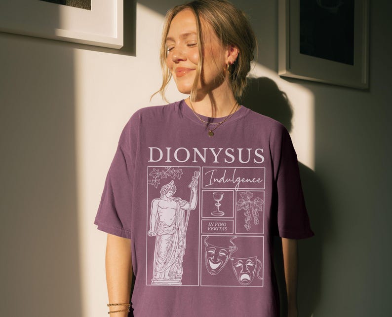 May include: A mauve-colored t-shirt with a graphic design featuring the word "DIONYSUS" and the word "Indulgence". The design includes a statue, a wine glass, grapes, and theatrical masks. The phrase "IN VINO VERITAS" is also included.