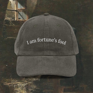 May include: Olive green corduroy baseball cap with the embroidered text "I am fortune's fool" in white. The cap has a curved brim and a top button. The hat is a classic style, suitable for casual wear.