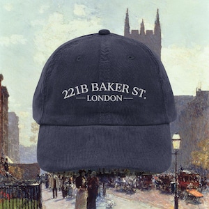 May include: Navy blue corduroy baseball cap with the text "221B BAKER ST. LONDON" in white. The cap has a curved brim and a rounded top. The background is a vintage painting of a street scene.