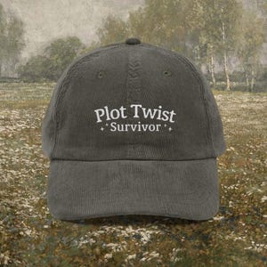 May include: A dark green corduroy baseball cap with the text "Plot Twist Survivor" embroidered in white. The cap has a curved brim and a rounded top. The background is a blurred image of a field and trees.
