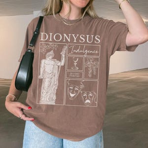 May include: Brown t-shirt with the text "DIONYSUS Indulgence" and illustrations of a statue, wine glass, grapes, and theater masks. The shirt is a solid brown color.