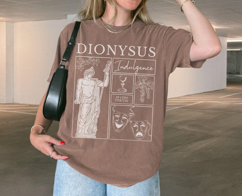 May include: Brown t-shirt with the text "DIONYSUS Indulgence" and illustrations of a statue, wine glass, grapes, and theater masks. The shirt is a solid brown color.
