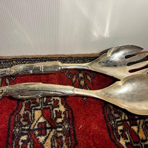 May include: A pair of silver-toned metal salad servers featuring handles moulded into the shape of birds. The top utensil is a three-pronged fork, while the bottom utensil is a large, rounded spoon. Both pieces have detailed, textured metalwork.