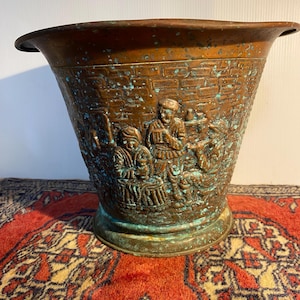 May include: A tapered copper bucket featuring a relief scene of three figures in a brick-walled setting. The metal surface displays a weathered brown finish with prominent teal-coloured oxidation spots throughout the exterior and rim.