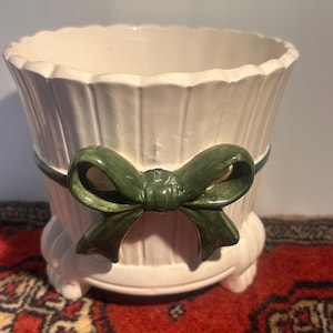 May include: A cream-coloured ceramic planter featuring a vertical ribbed texture and a scalloped rim. The exterior is decorated with a prominent, glossy dark green sculpted bow that wraps around the centre. The planter rests on small, rounded feet.