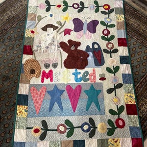 May include: A rectangular patchwork quilt featuring a cream-coloured centre with appliquéd designs including a brown teddy bear, a purple butterfly, hearts, stars, and the word "Metted" in colourful fabric letters, surrounded by a multi-coloured border.