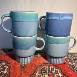 May include: Four ceramic mugs arranged in two stacks. Each mug features a two-tone blue glaze with a visible crackle finish on the lower section and a smooth, solid blue glaze on the upper rim. The bases of the bottom mugs have vertical stripes.