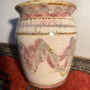 May include: A ceramic vase with a wide mouth and a textured surface. The vase features a cream-coloured base with pink, grey, and brown abstract patterns. The rim and base have a brown trim.